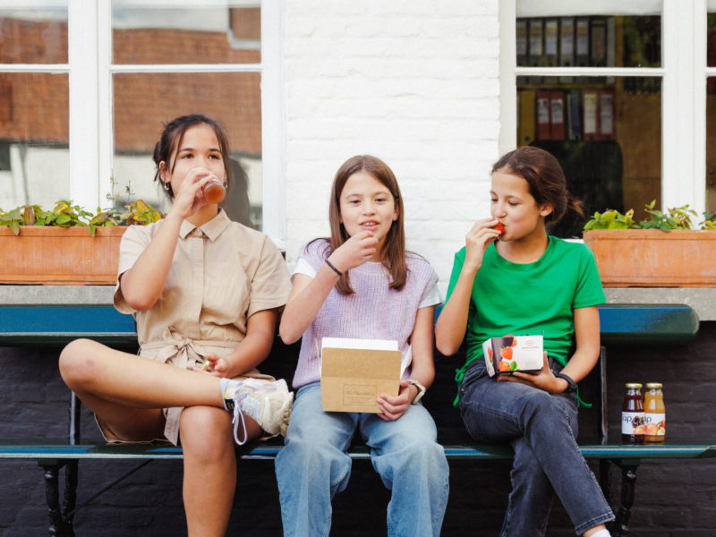 3 kinderen die samen op een bankje zitten met een vruchtensapje, een doosje aardbeien en een doosje pralines.